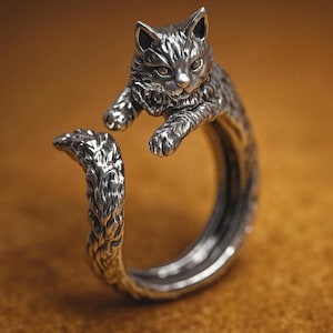 May include: A silver-toned ring featuring a detailed cat design. The cat's head and paws extend over the ring's band, with the tail wrapping around. The ring has a textured finish, giving it a vintage or handcrafted appearance.
