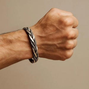 Puede incluir: Una pulsera trenzada de color plateado se lleva en la muñeca de una persona. La pulsera tiene un diseño grueso y texturizado con hebras entrelazadas. El fondo es de un color sólido y neutro, resaltando los detalles de la pulsera.