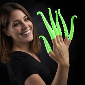 May include: A person's hand wearing five neon green tentacle finger covers. The covers are curved and have a textured surface with small, round bumps. The background is black.