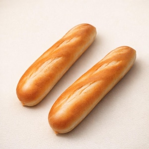 May include: Two golden-brown baguettes with a slightly textured surface. The bread has diagonal slashes across the top. The baguettes are displayed on a light-colored background.