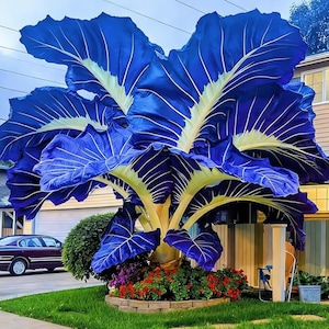 May include: A large, eye-catching sculpture of a plant with oversized blue leaves and yellow veins. The plant has a thick, yellow trunk and is surrounded by red flowers and green grass. A car and house are in the background.