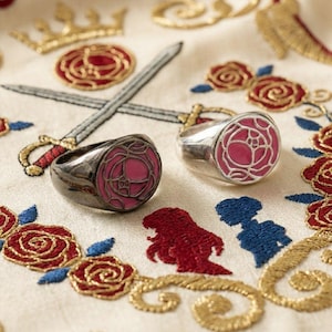 May include: Two signet rings featuring a circular face with a pink rose enamel inlay. One ring is crafted from a dark gunmetal-toned metal, while the other is made of a polished silver-toned metal, both showcasing a detailed rose design.