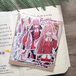 May include: A collection of die-cut vinyl stickers featuring the anime character Zero Two from Darling in the Franxx. The stickers display the character with long pink hair, red horns, and a red uniform, presented in a clear plastic sleeve.