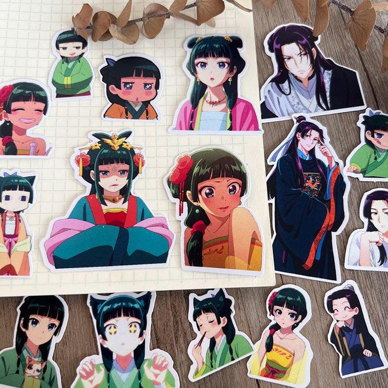 May include: A selection of anime-style stickers depicting characters in traditional Asian clothing. The stickers feature a variety of colours, including green, pink, and gold, with detailed facial expressions and hairstyles. The stickers are on a grid paper background.