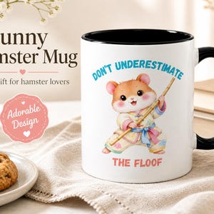 May include: A white ceramic mug with a black handle and interior rim. The front features a cartoon hamster wearing a light-coloured martial arts outfit, holding a wooden staff, with the text "DON'T UNDERESTIMATE THE FLOOF" printed in blue and red.