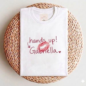 以下が含まれることがあります： 赤い文字で「hands up! Gabriella」と赤い唇のプリントが施された白いTシャツ。Tシャツは織りの丸いプレースマットの上に折りたたまれています。