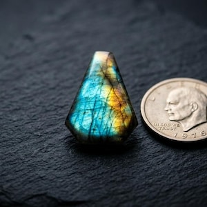 以下が含まれることがあります： 虹色の青と金の輝きを持つ、磨かれた涙型のラブラドライトの宝石。隣には1976年の銀貨。宝石は暗く、質感のある表面に展示されています。