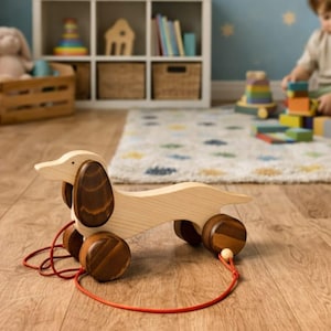 Può includere: Giocattolo a forma di bassotto in legno da tirare, con orecchie e ruote marroni. Il giocattolo ha un cordino rosso ed è realizzato in legno chiaro. Un giocattolo classico per bambini piccoli.