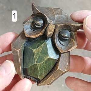Può includere: Una statuetta di gufo in legno intagliato con un design geometrico. Il gufo ha tonalità marrone scuro e verde, con intagli dettagliati sulle piume e sugli occhi. Il numero "1" è nell'angolo in alto a sinistra.