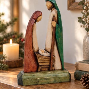 Könnte beinhalten: Eine geschnitzte Krippenfigur aus Holz mit zwei Personen, die über einer kleinen Krippe stehen. Eine Figur trägt einen dunkelroten Umhang, die andere einen grünen Umhang, beide mit cremefarbenen Gewändern. Sie stehen auf einem grünen Sockel.