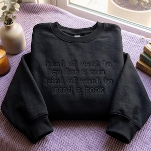 Puede incluir: Una sudadera negra de cuello redondo con bordado negro tonal en el pecho con el texto: kind of want to go for a run kind of want to read a book. La prenda tiene mangas largas con puños acanalados y un cuello también acanalado.