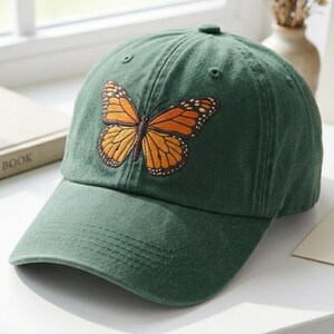 Può includere: Un cappellino da baseball verde con un ricamo di farfalla monarca. La farfalla ha ali arancioni e marroni con dettagli in bianco e nero. Il cappellino ha una visiera curva e un bottone sulla parte superiore.