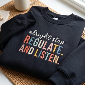 Op de afbeelding: Een marineblauwe sweatshirt met de geborduurde tekst "alright stop REGULATE. AND LISTEN." De tekst is veelkleurig. Op de achtergrond is een beige mok te zien. De sweatshirt ligt op een gevlochten mat.