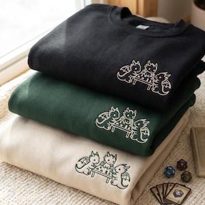 May include: A stack of three crewneck sweatshirts in black, dark green, and cream. Each sweatshirt features an embroidered design on the chest showing three small dragons sitting around a square table playing a board game with dice.