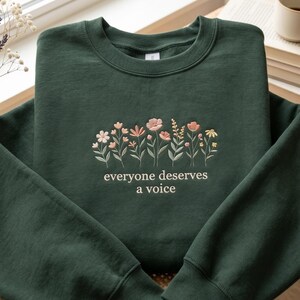 以下が含まれることがあります： ピンク、ピーチ、イエローの色合いの刺繍の花柄が施された濃い緑色のスウェットシャツ。「everyone deserves a voice」の文字が花の下に刺繍されています。スウェットシャツは明るい色の表面に折りたたまれています。