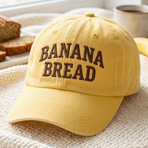 May include: A yellow baseball cap with the words "BANANA BREAD" embroidered in brown. The cap has a curved peak and a button on top. The hat is on a white, textured surface.