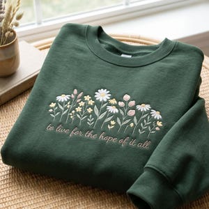 Op de afbeelding: Een donker bosgroene sweater met ronde hals voorzien van een borduursel van diverse wilde bloemen in witte, gele en zachtroze tinten. Onder het bloemmotief is de tekst "to live for the hope of it all" geborduurd met lichtroze garen.