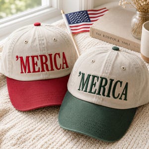 Könnte beinhalten: Zwei Baseballkappen mit beigen Kronen und gesticktem Text 'MERICA. Eine Kappe hat einen roten Schirm und einen roten Knopf oben, während die andere einen dunkelgrünen Schirm und einen dunkelgrünen Knopf oben hat. Beide haben sechs Paneele.