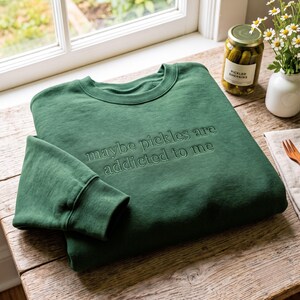 May include: A forest green crewneck sweatshirt featuring the embroidered text "maybe pickles are addicted to me" in a matching dark green thread. The long-sleeved garment has a ribbed collar, cuffs, and hem, showcasing a simple, solid-coloured design.
