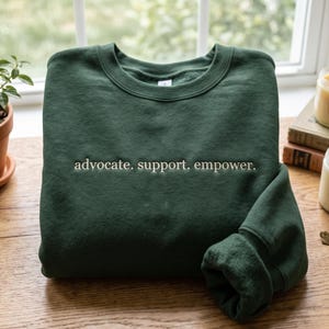 Könnte beinhalten: Ein dunkelwaldgrünes Sweatshirt mit Rundhalsausschnitt und dem gestickten Text "advocate. support. empower." in hellcremefarbenem Garn, mittig auf der Brust platziert. Das Kleidungsstück hat lange Ärmel sowie gerippte Bündchen am Kragen, an den Ärmeln und am Saum.