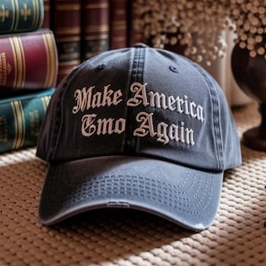 May include: A distressed navy blue baseball cap with the embroidered phrase "Make America Emo Again" in white script. The cap has a curved brim and a worn appearance, suggesting a vintage or retro style.