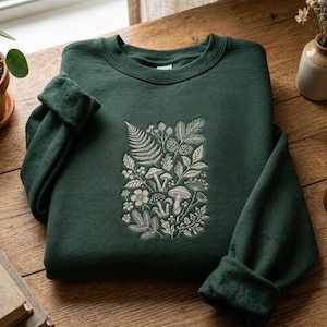 May include: A dark forest green crewneck sweatshirt featuring a central embroidered design in light beige thread. The embroidery depicts a detailed botanical arrangement of various mushrooms, fern fronds, pine cones, and small leafy sprigs.