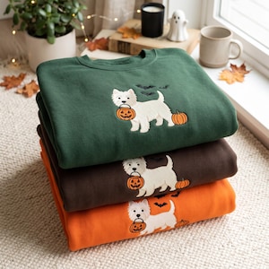 May include: A stack of three crewneck sweatshirts in dark green, dark brown, and orange. Each sweatshirt features an embroidered white West Highland White Terrier dog holding an orange jack-o'-lantern bucket, with small black bats and a pumpkin.