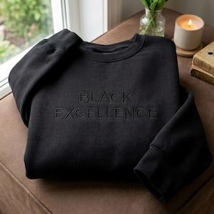 Puede incluir: Una sudadera negra de manga larga con cuello redondo que presenta las palabras BLACK EXCELLENCE bordadas en hilo negro a juego en el centro del pecho. La prenda tiene cuello, puños y dobladillo acanalados con un acabado negro sólido.