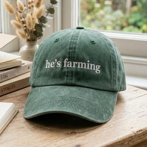 May include: A green baseball cap with the embroidered text "he's farming" in white. The hat is made of a soft fabric and has a curved brim. The hat is sitting on a wooden surface.