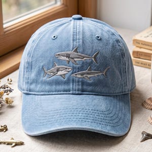 May include: A light blue denim baseball cap featuring an embroidered design of three grey sharks swimming in a group. The cap has a structured crown, a curved brim, and stitched eyelets on the top panels for ventilation and a classic athletic look.
