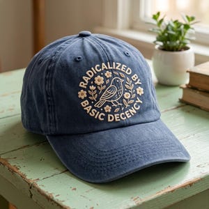 Op de afbeelding: Een marineblauwe baseballcap met de geborduurde tekst "RADICALIZED BY BASIC DECENCY" in een cirkelvormig ontwerp. Het ontwerp bevat een vogel en bloemmotieven in crème. De cap ligt op een verweerde groene tafel.