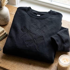 May include: A black crewneck sweatshirt featuring three embroidered dragonflies on the front. The embroidery is tonal, matching the black fabric of the garment. The sweatshirt has long sleeves and a ribbed collar, cuffs, and hemline.