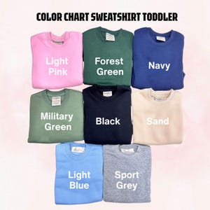 May include: A color chart for toddler sweatshirts featuring eight crewneck pullovers in light pink, forest green, navy, military green, black, sand, light blue, and sport grey. Each sweatshirt is labeled with its corresponding color name in white text.