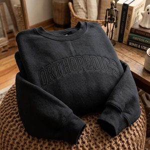 May include: A black long-sleeve crewneck sweatshirt featuring the word ORTHOPEDICS embroidered in a matching black thread across the chest in a bold, arched collegiate-style font. The garment has ribbed cuffs and a ribbed neckline.