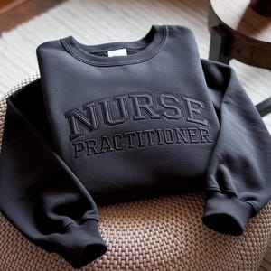 Könnte beinhalten: Ein dunkelgraues Sweatshirt mit dem gestickten Schriftzug "NURSE PRACTITIONER" auf der Brust. Das Sweatshirt ist gefaltet und liegt auf einer gewebten Oberfläche. Die Ärmel haben gerippte Bündchen.