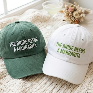 Może przedstawiać: Dwie czapki bejsbolowe, jedna zielona i jedna biała, z wyhaftowanym napisem "THE BRIDE NEEDS A MARGARITA". Zielona czapka ma biały napis, a biała czapka ma zielony napis. Czapki leżą na kremowej, teksturowanej powierzchni.