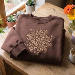 May include: A dark brown crewneck sweatshirt featuring a large, intricate, light-colored floral mandala embroidery centered on the chest. The garment has long sleeves with ribbed cuffs and a ribbed neckline, showcasing a detailed botanical pattern.