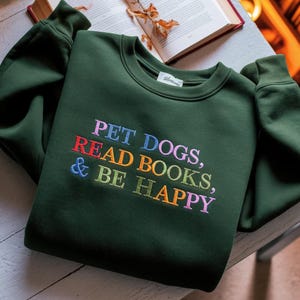 May include: A dark green sweatshirt with the embroidered text "PET DOGS, READ BOOKS, & BE HAPPY" in colorful letters. The sweatshirt has a crew neck and long sleeves. The image also shows a partially visible open book.