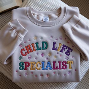 May include: A beige sweatshirt with the words "CHILD LIFE SPECIALIST" embroidered in colorful letters. The design includes embroidered starbursts around the text. The sweatshirt is folded neatly on a textured surface.