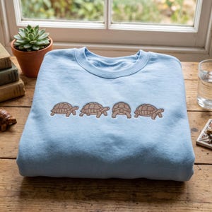 May include: A light blue crewneck sweatshirt featuring an embroidered design of four brown tortoises arranged in a horizontal row across the centre. The tortoises are depicted in various walking positions, with one facing forward and three facing sideways.