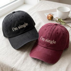 May include: Two baseball caps, one black and one maroon, each featuring the embroidered text "i'm single" in white on the front panel. Both caps have a structured crown, a curved brim, and a button at the top, showcasing a classic six-panel design.