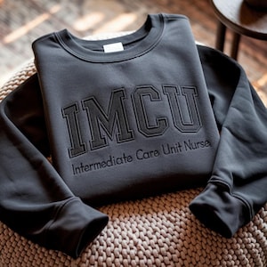 May include: A dark gray sweatshirt with the embroidered letters "IMCU" above the text "Intermediate Care Unit Nurse". The sweatshirt is folded on a woven surface.