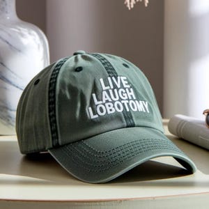 May include: A khaki green baseball cap with the slogan "LIVE LAUGH LOBOTOMY" in white capital letters. The cap features a curved peak and stitched detailing. It is displayed on a light-coloured surface.