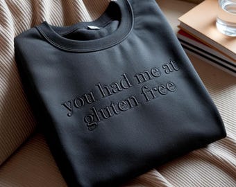 May include: A charcoal grey crewneck sweatshirt featuring the embroidered text you had me at gluten free in a matching dark thread. The garment has a ribbed collar and long sleeves, showcasing a clean, monochromatic design with textured lettering.