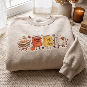 Puede incluir: Una sudadera de cuello redondo en color beige con un bordado de libros antropomórficos. El diseño incluye una pila de libros con una taza de café, un libro rojo con el texto PUMPKIN TALES, un libro amarillo con COZY CLASSICS y un libro abierto.