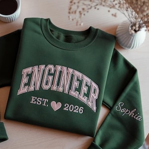 Könnte beinhalten: Ein dunkelgrüner Rundhals-Pullover mit hellrosa Stickerei, die ENGINEER EST. 2026 mit einem Herzsymbol in der Mitte zeigt. Auf dem Ärmel ist der Name Sophia in passender hellrosa Schreibschrift eingestickt.