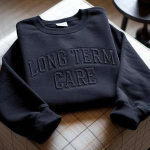 以下が含まれることがあります： 黒色のクルーネックのスウェットシャツ。胸には「LONG TERM CARE」の文字が同系色の濃い色で刺繍されています。スウェットシャツは模様のある表面に折りたたまれています。
