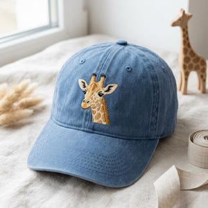 Puede incluir: Una gorra de béisbol azul con una cabeza de jirafa bordada en el panel frontal. La gorra tiene una visera curva, un botón en la parte superior y ojales cosidos para la ventilación. El bordado de la jirafa muestra manchas marrones sobre fondo beige.