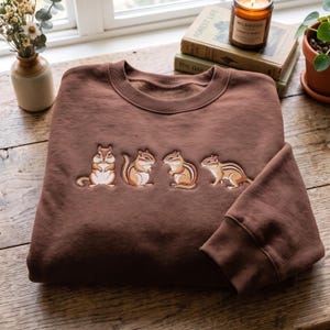 Peut inclure: Un sweat-shirt marron à col rond orné d'une broderie représentant quatre tamias dans des poses variées sur la poitrine. Le vêtement possède des manches longues avec des poignets côtelés et un col côtelé, fabriqué en tissu marron uni.