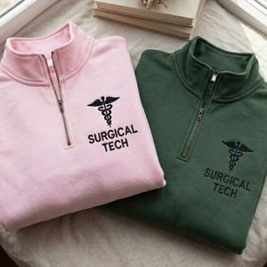 Può includere: Due pullover con zip corta, uno rosa e uno verde. Ognuno presenta un simbolo medico nero ricamato e la scritta "SURGICAL TECH". I pullover sono piegati su una superficie chiara.